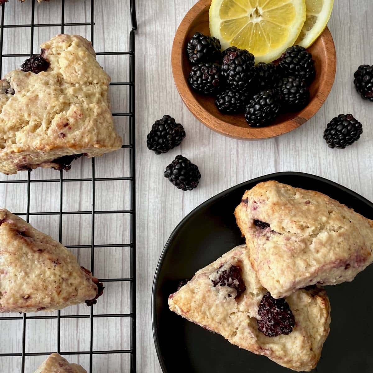 Baked scones on a cooling rack and plate next to fresh lemons and blackberries.